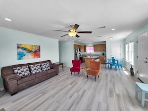 A spacious living room with a brown sofa, red and orange chairs, a ceiling fan, and a kitchen in the background.