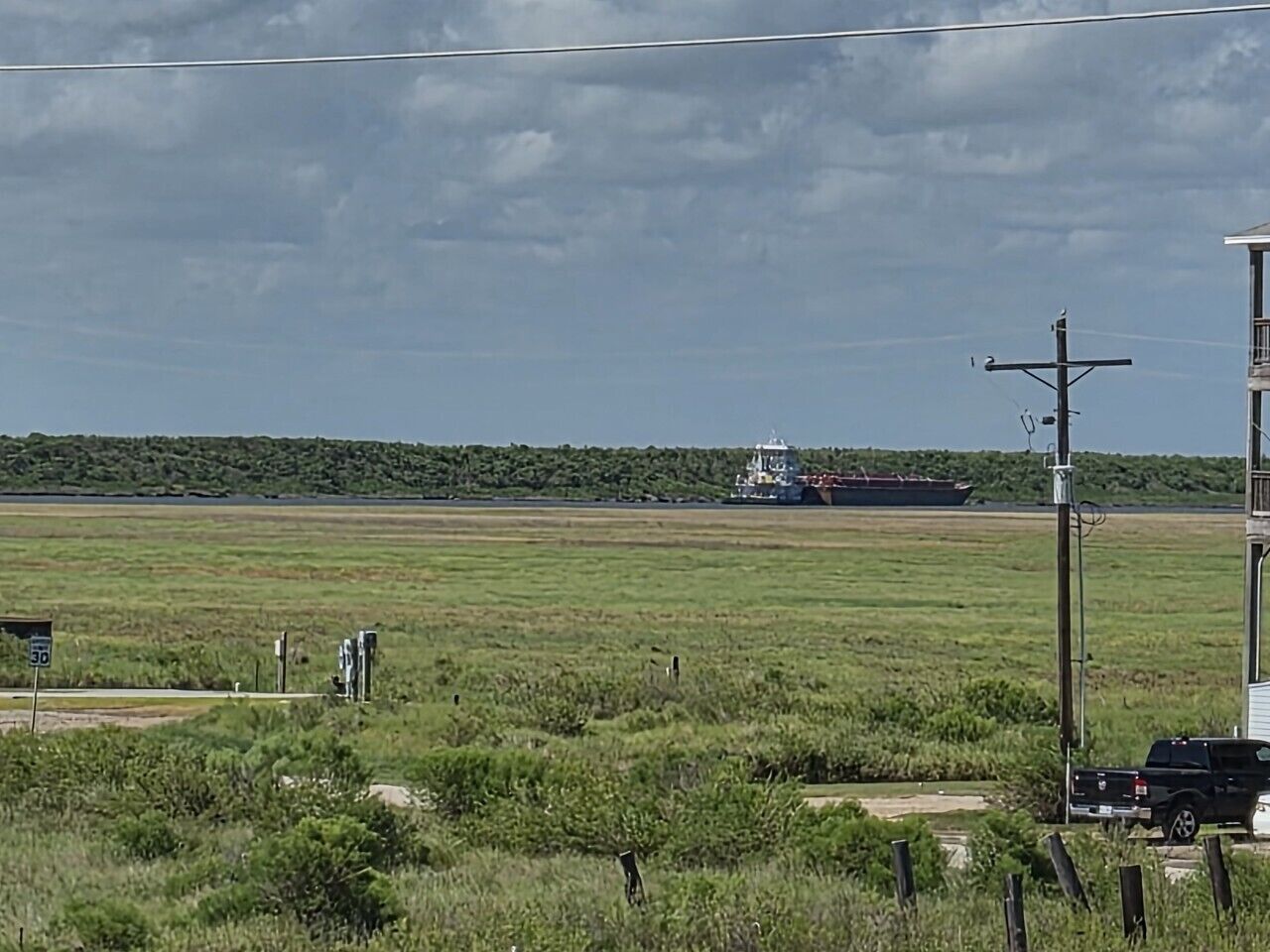 A grassy field with a large ship in the distance, a truck, and a building with a balcony.