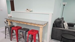 A room with a light blue wall, a wooden bench with a granite countertop, three red stools, a grey sofa, and a black table. - Crabby Cove - Fully Fenced Side Yard, Pets OK, Gulf & Bay Views, Walk to Beach (Bolivar Peninsula)