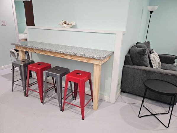 A room with a light blue wall, a wooden bench with a granite countertop, three red stools, a grey sofa, and a black table.