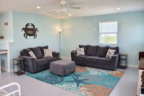 A living room with two grey sofas, a matching ottoman, a patterned rug, a ceiling fan, and a window with blinds.