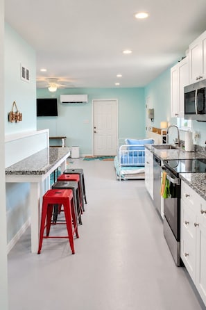 Une cuisine moderne avec un îlot central, un coin repas avec des tabourets rouges et une chambre avec un lit bleu.