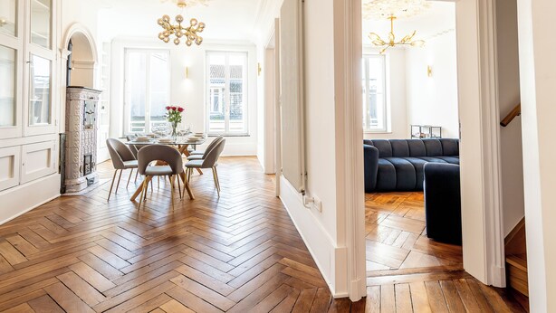 A spacious room with a herringbone floor, a dining area with a table and chairs, a black sofa, and a chandelier.