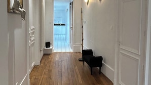 A hallway with wooden flooring, white walls, and a black chair. There is a door on the right and a framed sculpture on the left.