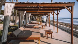 Un salon de plage avec des chaises longues et des tables en bois, une pergola dotée d’un toit en tuiles et une vue sur l’océan.