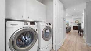A laundry room with two Samsung washing machines, a white cabinet, and a dining area with a table and chairs. - Sandcastle by Ryson Vacation Rentals (Galveston)