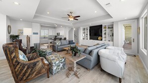 A spacious living room with a high ceiling, a ceiling fan, and a large TV. There is a kitchen area in the background with white cabinets and a dining table. - Sandcastle by Ryson Vacation Rentals (Galveston)