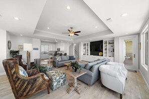 A spacious living room with a high ceiling, a ceiling fan, and a large TV. There is a kitchen area in the background with white cabinets and a dining table. - Sandcastle by Ryson Vacation Rentals (Galveston)