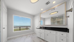 A spacious bathroom with a large mirror, white cabinetry, and a view of the ocean. - Sandcastle by Ryson Vacation Rentals (Galveston)