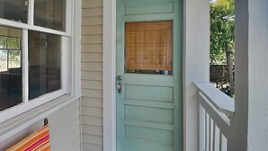 Uma porta da frente com porta verde e janela com persianas, um alpendre com um banco listrado, e uma cerca de madeira ao fundo.