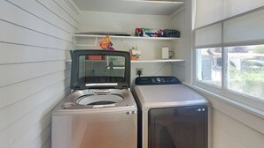 Uma área de lavandaria com máquina de lavar de abertura frontal e secadora, uma janela com vista para o exterior e prateleiras com diversos objetos.