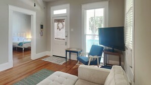 Un salon avec un canapé, un téléviseur et un fauteuil. On y trouve une porte avec une fenêtre et une petite table.
