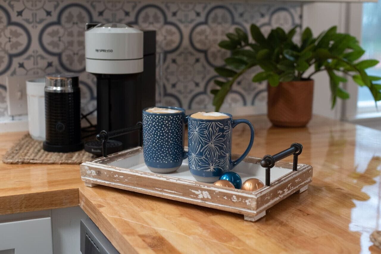 Una encimera de cocina con cafetera, dos tazas con diseños, una bandeja con adornos y una planta en maceta.