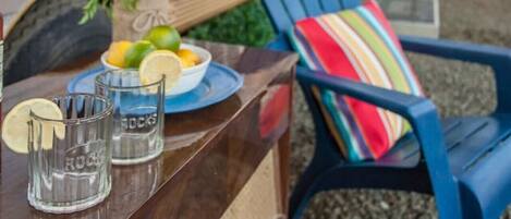 A wooden table with a plate of food, glasses, and a chair with a colorful cushion.