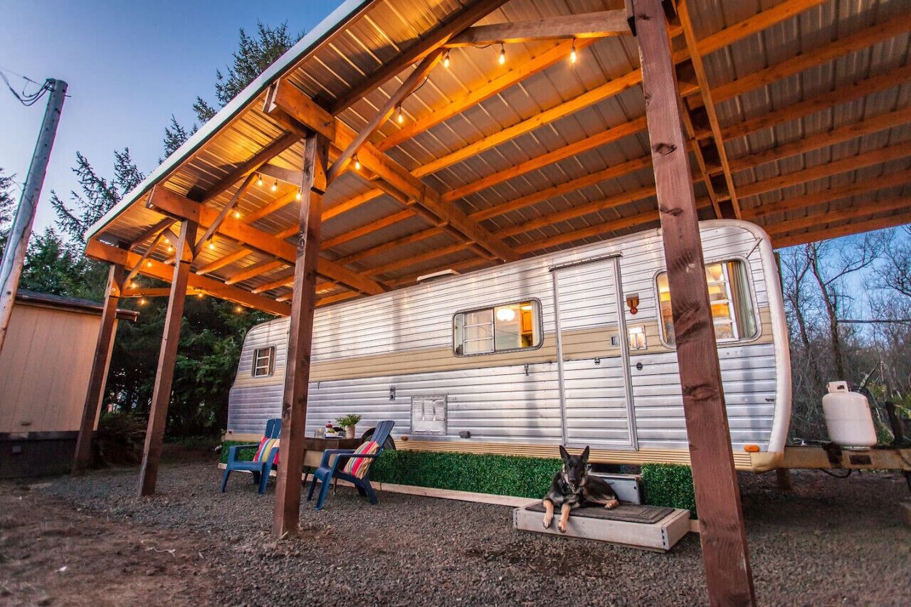 Un terrain de camping pour véhicules récréatifs, partiellement abrité, où un camping-car est garé sous une structure en bois.