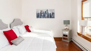 A hotel room with a bed, pillows, a nightstand with a lamp, a cityscape painting, and a window with blinds.