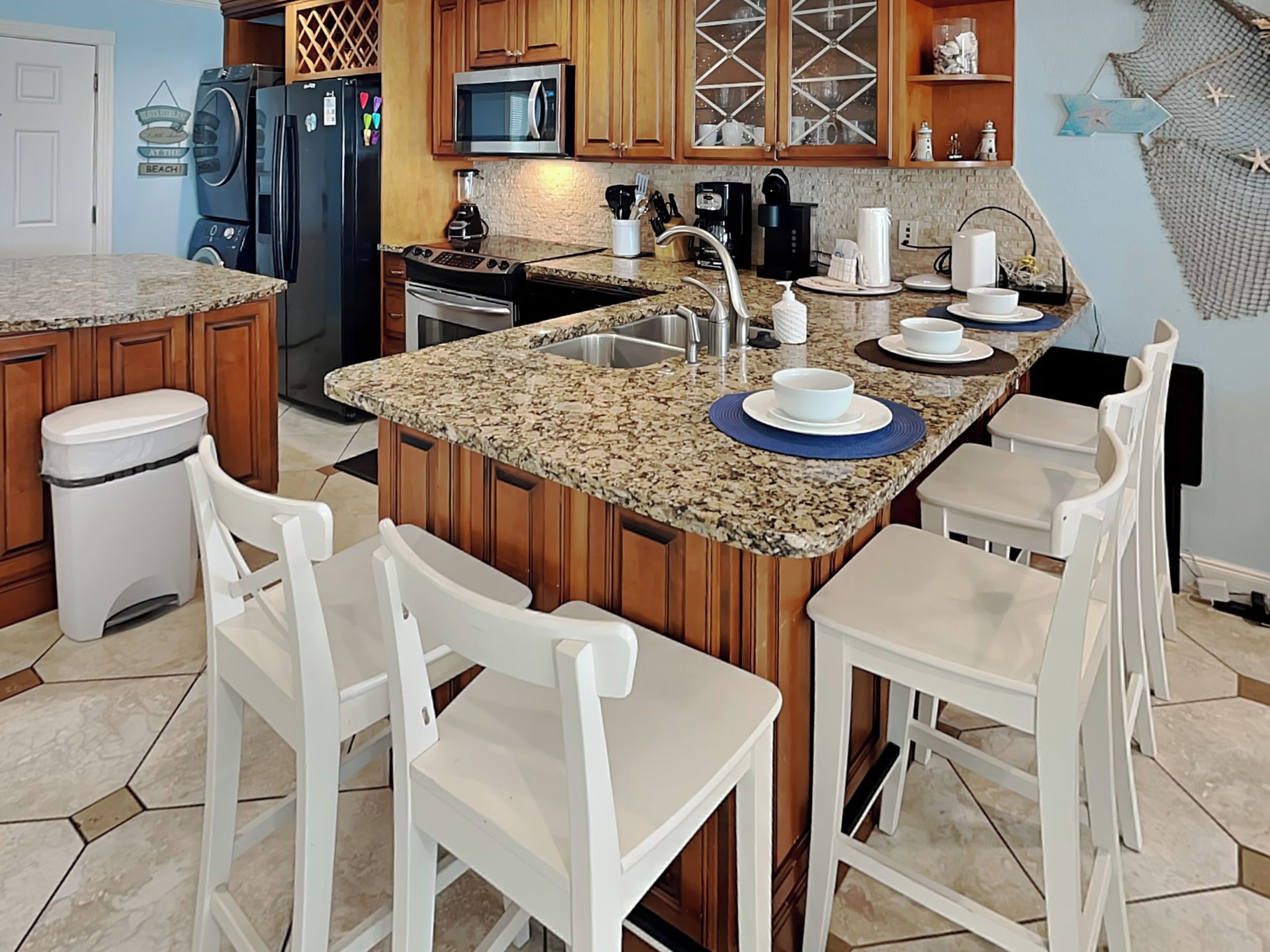 A kitchen with granite countertops, wooden cabinets, a stainless steel refrigerator, a microwave, and a sink. There are four white bar stools and a coffee maker on the counter.