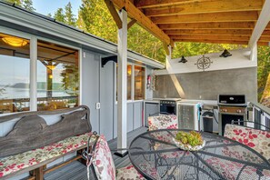 A covered outdoor patio with a wooden ceiling, a round metal table with a floral tablecloth, a bench with a floral cushion, and a grill station. - Hood Canal Views: Remote Cabin in Quilcene! (Quilcene)