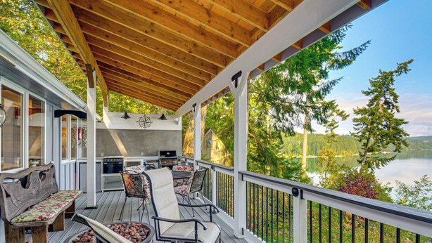 Une terrasse couverte avec coin salon extérieur, un barbecue et une vue sur un lac entouré d’arbres.