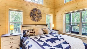 Un dormitorio de madera con una cama grande, dos mesitas de noche, un reloj en la pared y ventanas amplias con vistas a árboles.
