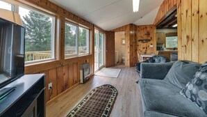Ein Blockhaus mit Holzbau, ein Wohnbereich mit grauem Sofa, Fernseher und eine durchgehende offene Tür zur Küche.