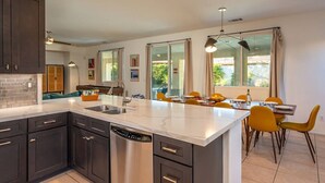 A modern kitchen with dark cabinets, a large island, and a dining area with yellow chairs. - Luma House | Stylish Getaway w/ Game Room & Pool[Free Early Check-In OR Late Checkout] (Indio)