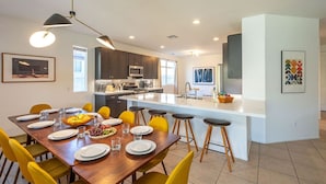 A modern kitchen with dark cabinets, a central island, and a dining area with yellow chairs and a wooden table. - Luma House | Stylish Getaway w/ Game Room & Pool[Free Early Check-In OR Late Checkout] (Indio)