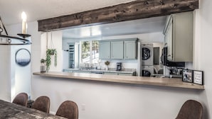 Un comedor con una mesa y sillas de madera, una lámpara colgante y vista a la cocina con armarios y electrodomésticos.