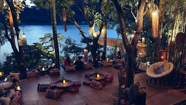 An outdoor seating area with round tables and cushioned chairs, surrounded by trees and overlooking a body of water. - Villa Lakangthong2 (Luang Prabang)