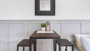 Un coin repas avec une table et des bancs, un miroir décoratif, une plante en pot et une lanterne sur un mur blanc avec des panneaux.