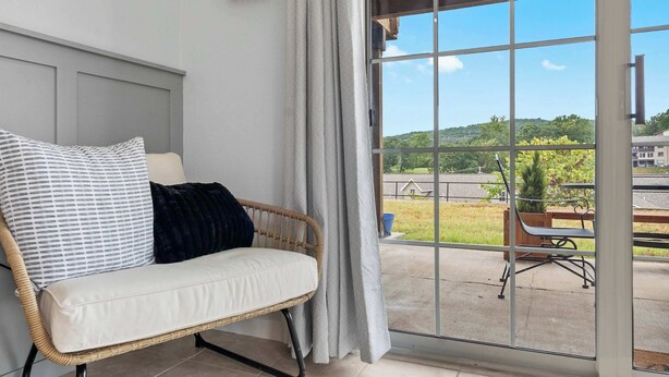 Un canapé en osier avec un coussin blanc et un oreiller noir, placé près d’une porte-fenêtre coulissante donnant sur un paysage verdoyant et un bâtiment situé au loin.