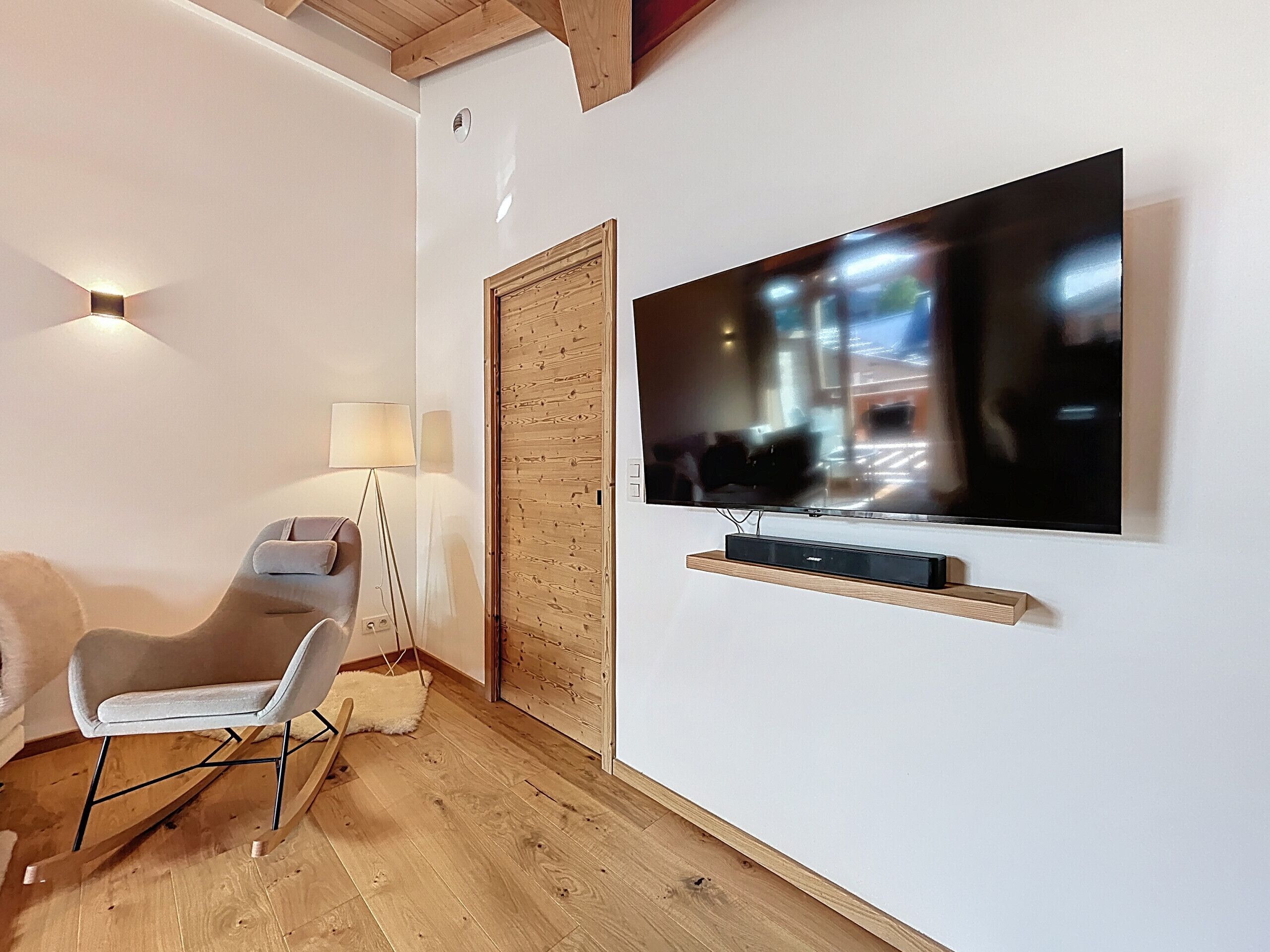 A modern living room with a flat-screen TV mounted on the wall, a grey armchair, a floor lamp, and a wooden door.