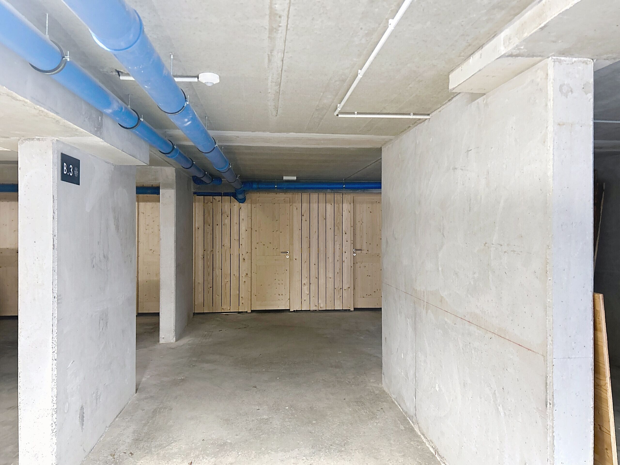 Interior de un estacionamiento con paredes de concreto, tuberías azules y una puerta de madera.