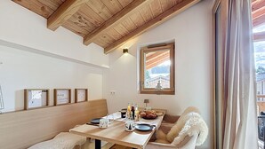 Un coin repas avec une table en bois, des chaises et un banc. Une fenêtre offre une vue sur un immeuble et un arbre.