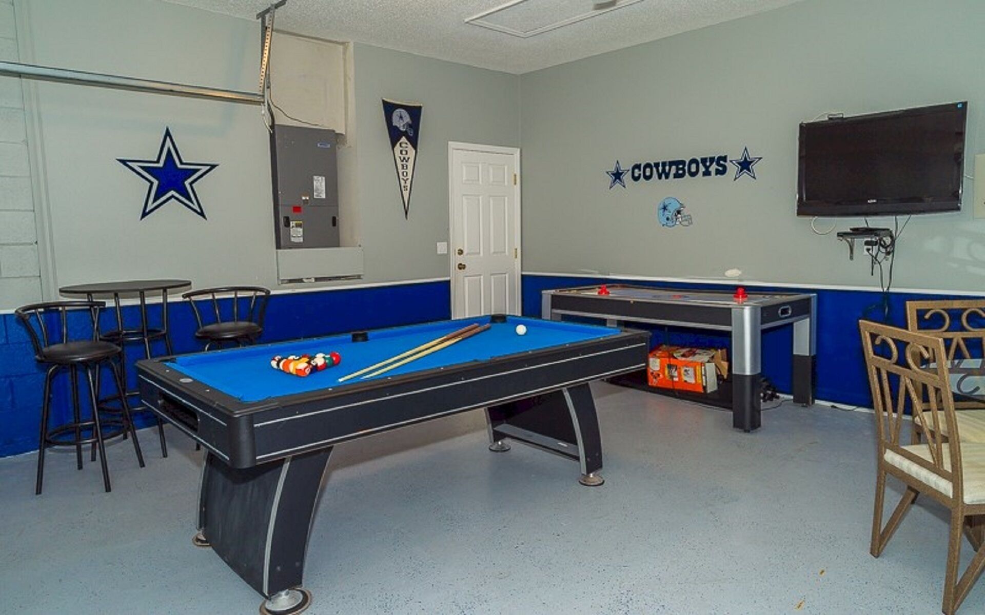 Une table de billard recouverte d’un drap bleu, un queue de billard et plusieurs billes. On y trouve également deux petites tables avec des chaises, une porte et un téléviseur fixé au mur.
