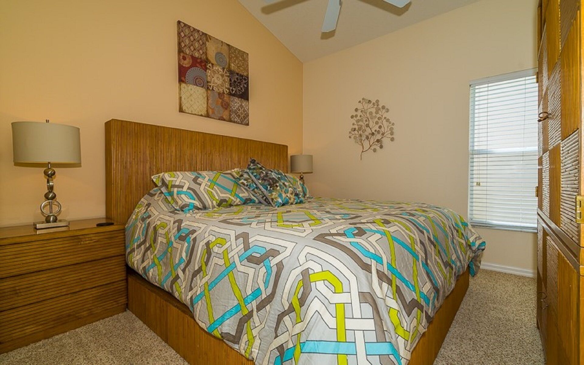 A bedroom with a bed, two bedside tables, a ceiling fan, a window with blinds, and a wall decoration.