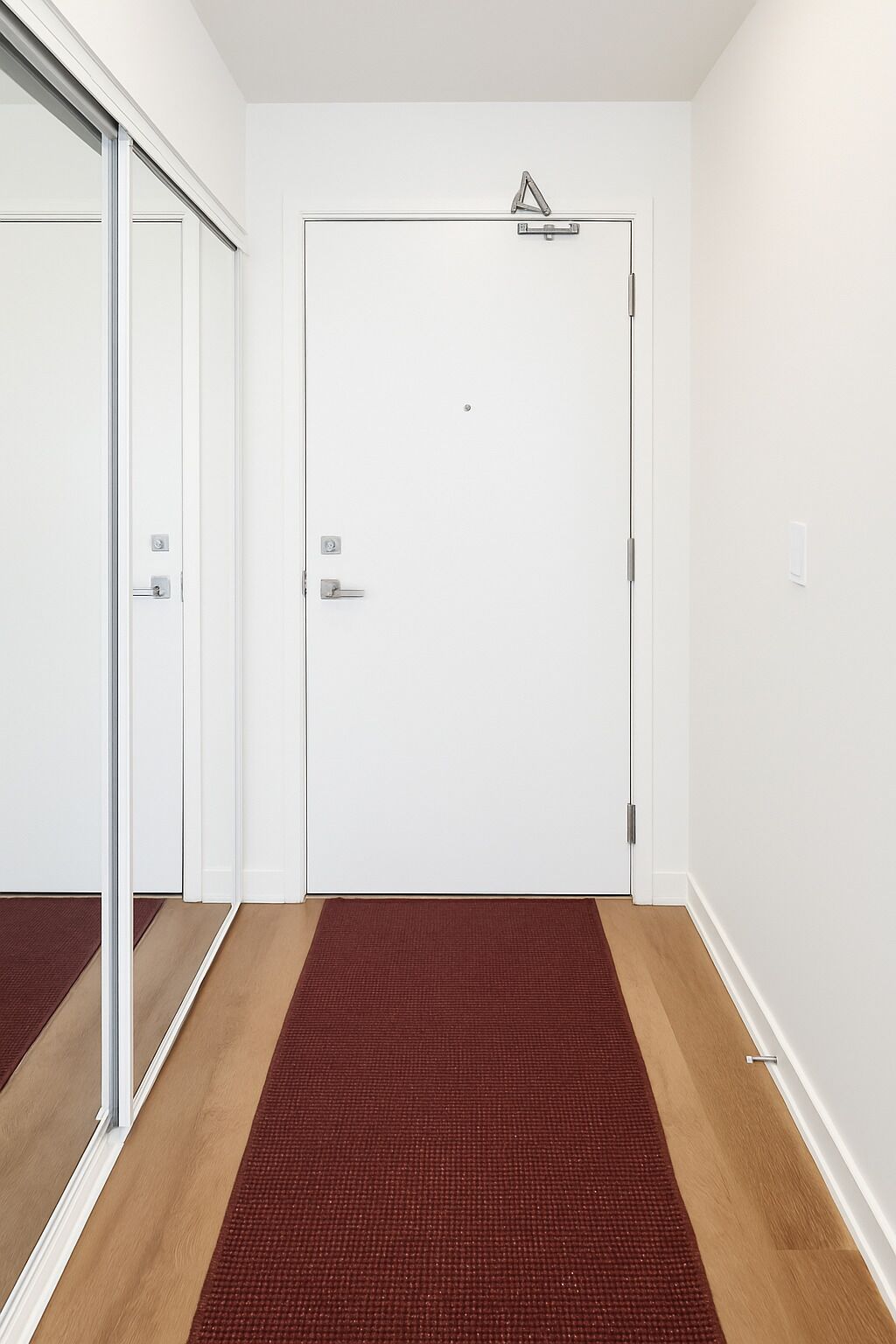 Una porta bianca con maniglia argentata, un tappetino rosso con trama decorativa all'ingresso e un piccolo cartello triangolare sopra la porta.