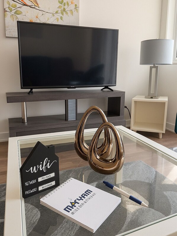 A modern living room with a flat-screen TV, a glass coffee table with a notebook, pen, and a decorative item.