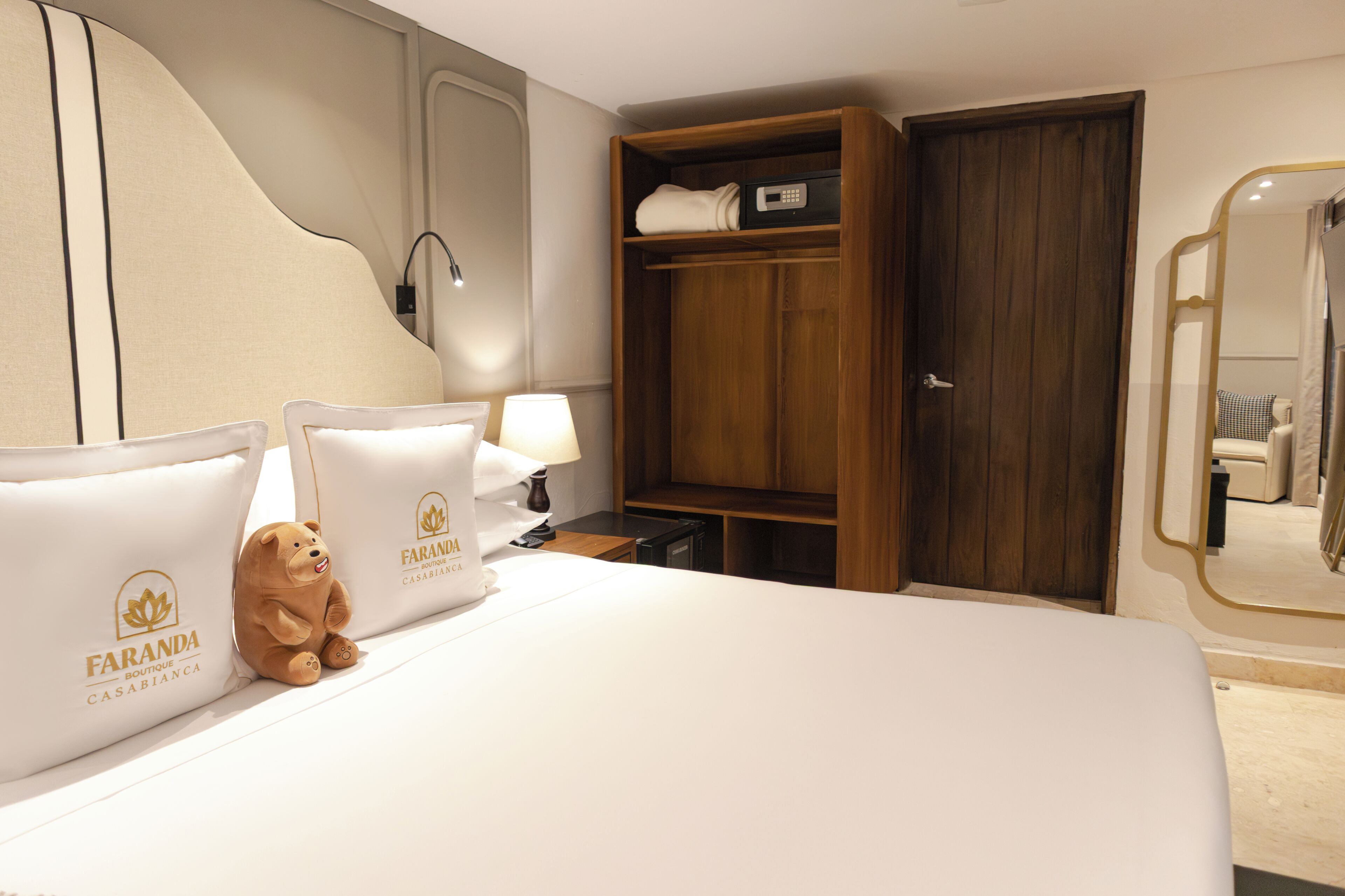 A hotel room with a bed, pillows, a nightstand, a lamp, a wooden wardrobe, and a teddy bear.