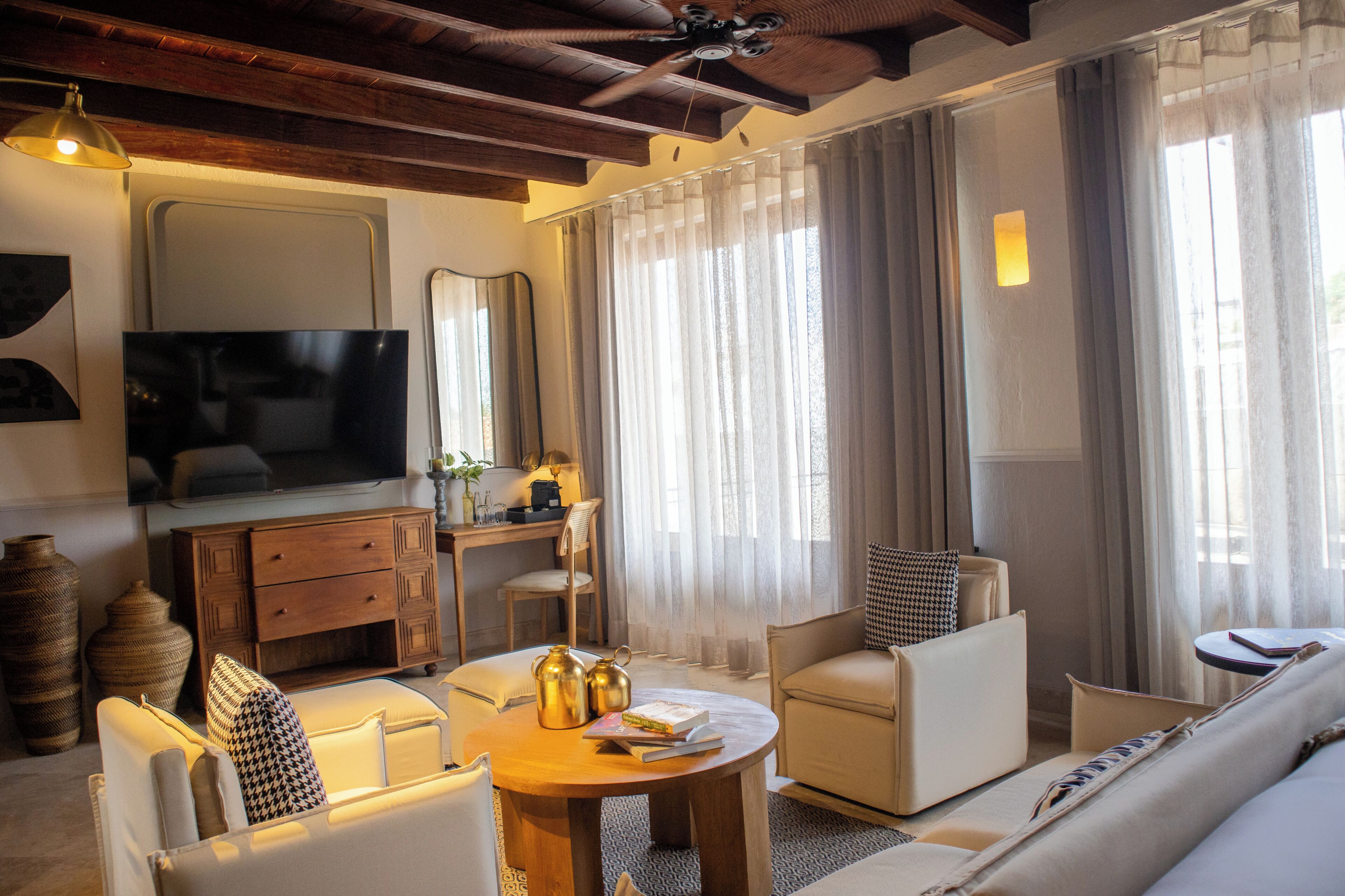 Uma sala de estar moderna com televisão de tela plana, móveis de madeira e grandes janelas com cortinas leves.