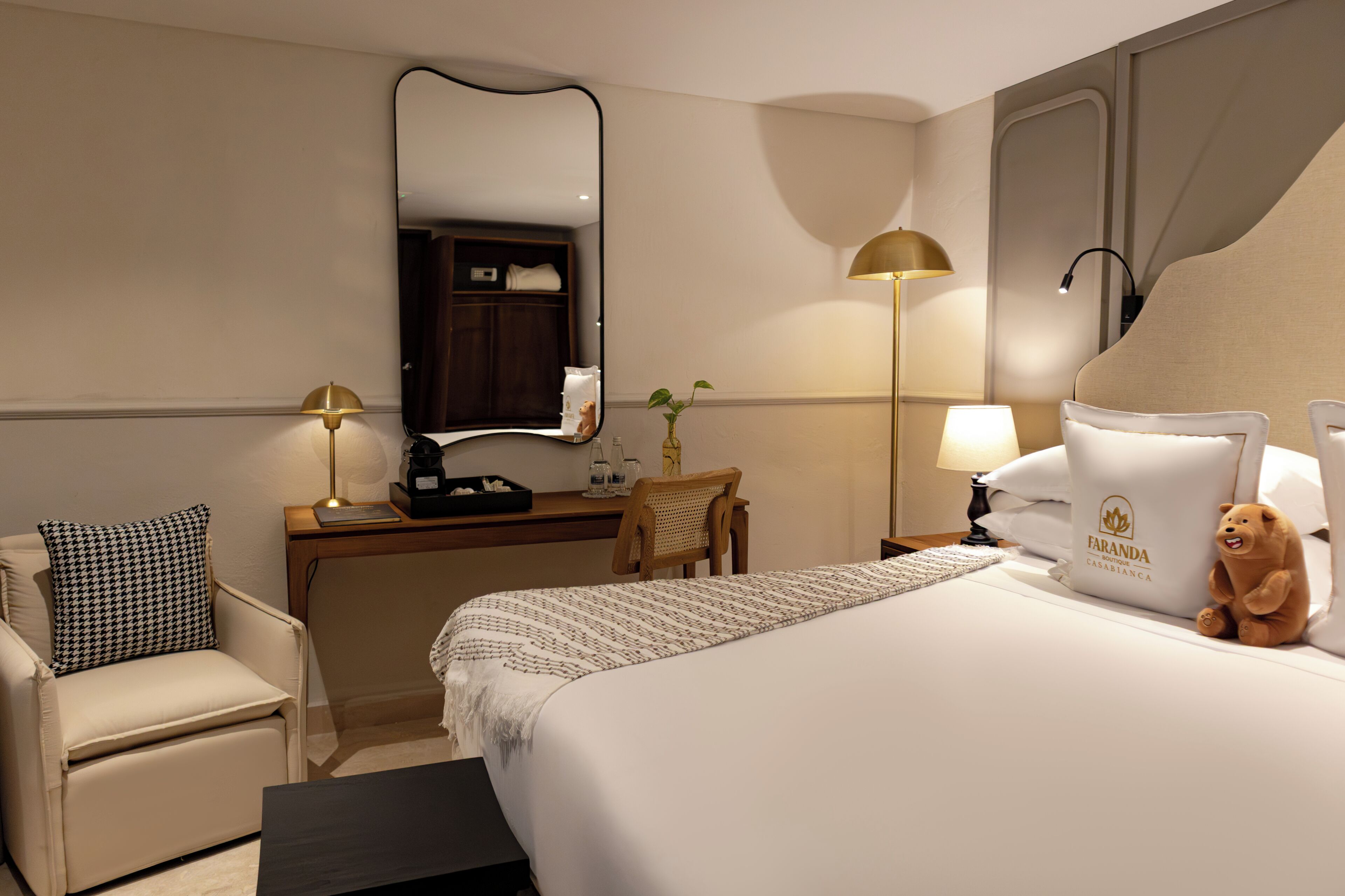 Une chambre d’hôtel avec un lit, une chaise, un bureau, une lampe, un ours en peluche et un miroir.
