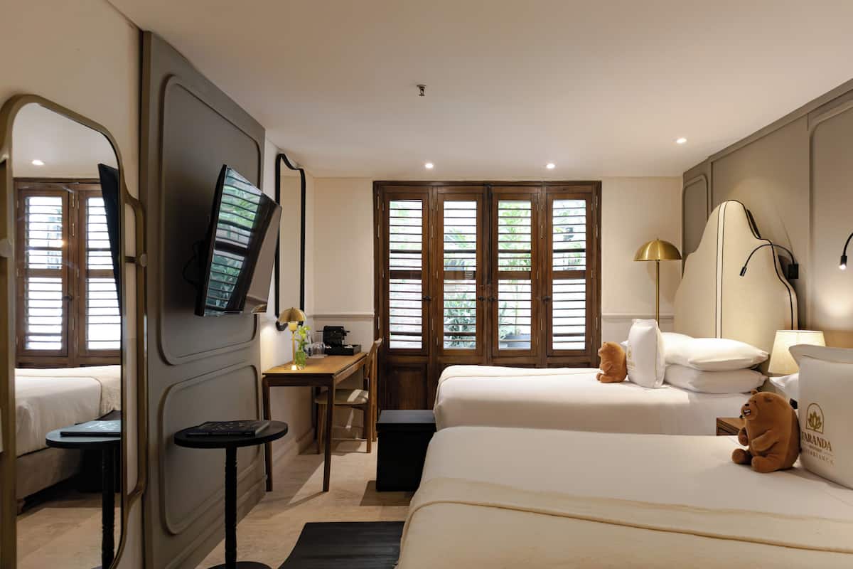 Habitación de hotel con cama, escritorio, silla, televisor y ventana con persianas.