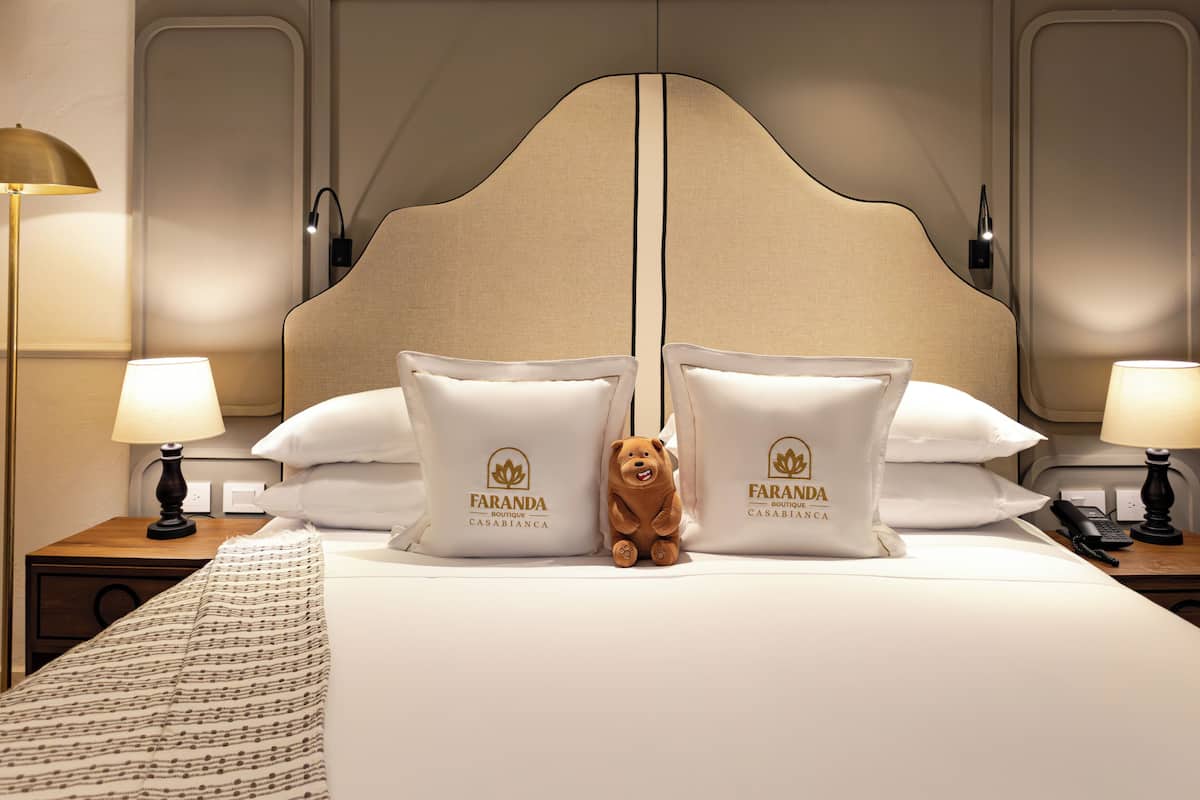 Habitación de hotel con una cama grande, dos mesitas de noche con lámparas, un osito de peluche y almohadas con el logotipo del hotel.