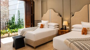 A hotel room with two beds, a small table, a nightstand, a lamp, and a view of greenery through a window. - Hotel Casa Bianca By Faranda Boutique (Cartagena)