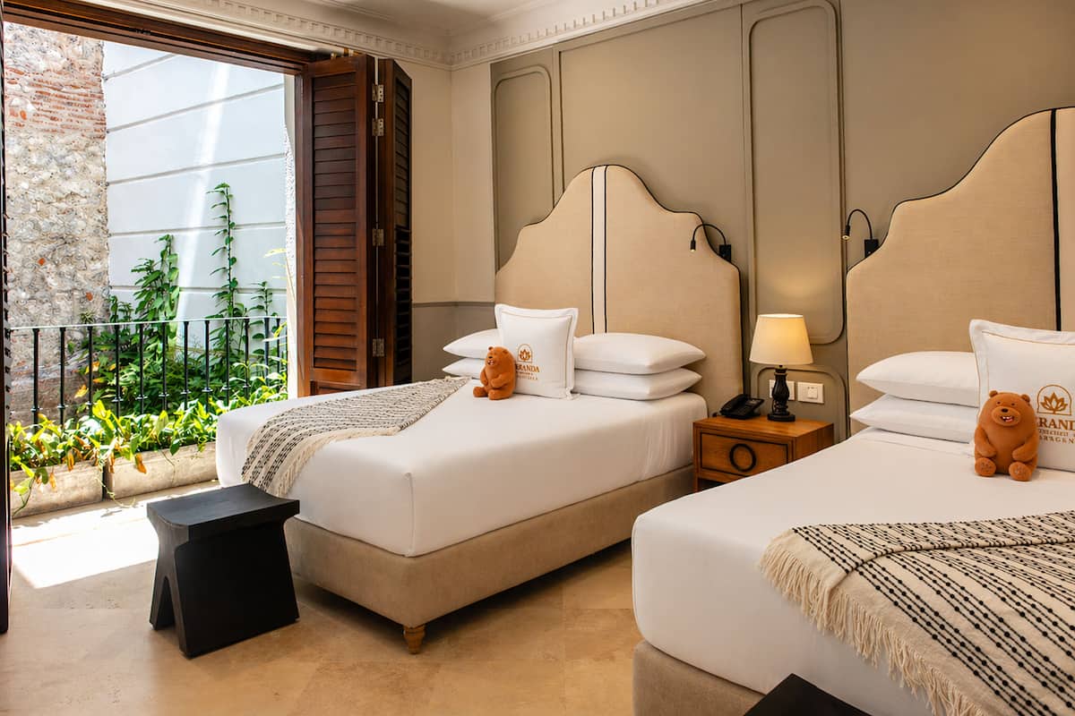 Habitación de hotel con dos camas, una mesita pequeña, una mesita de noche, una lámpara y vistas a la vegetación por la ventana.