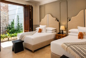 A hotel room with two beds, a small table, a nightstand, a lamp, and a view of greenery through a window.