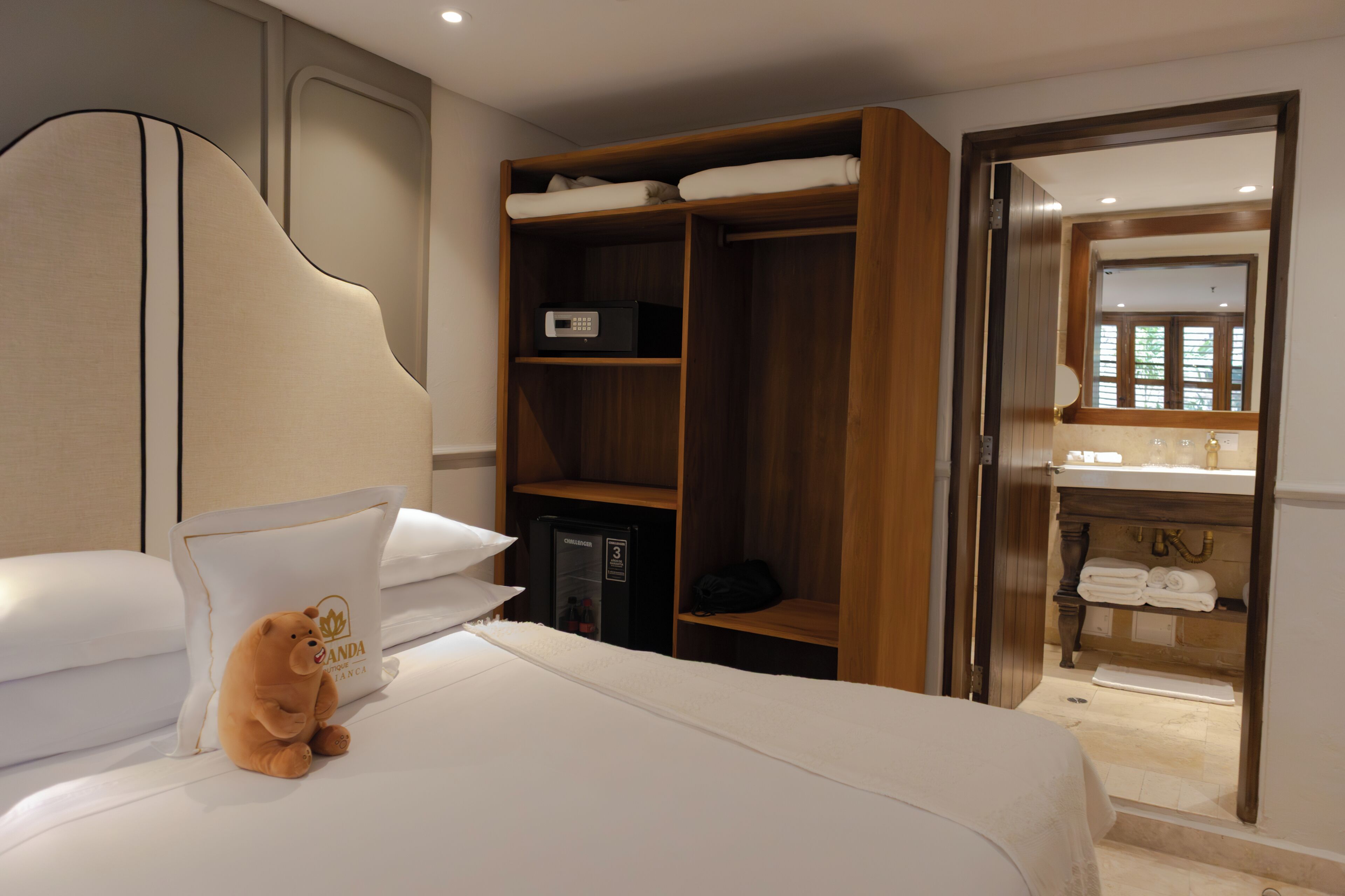 A hotel room with a bed, a teddy bear, a minibar, a wooden wardrobe, and a bathroom visible through an open door.