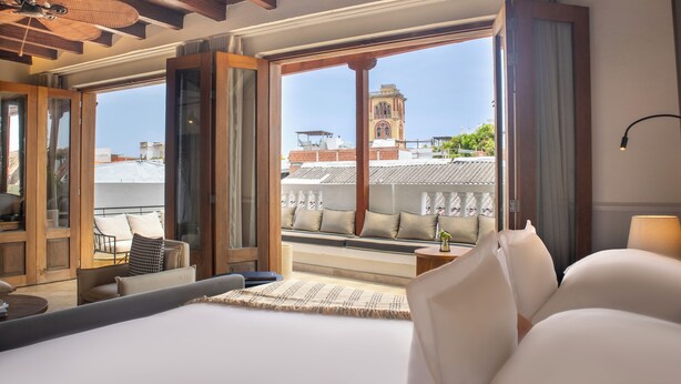 A hotel room with a large bed, a seating area with cushions, a view of a tower, and a balcony with a railing. - Hotel Casa Bianca By Faranda Boutique (Cartagena)
