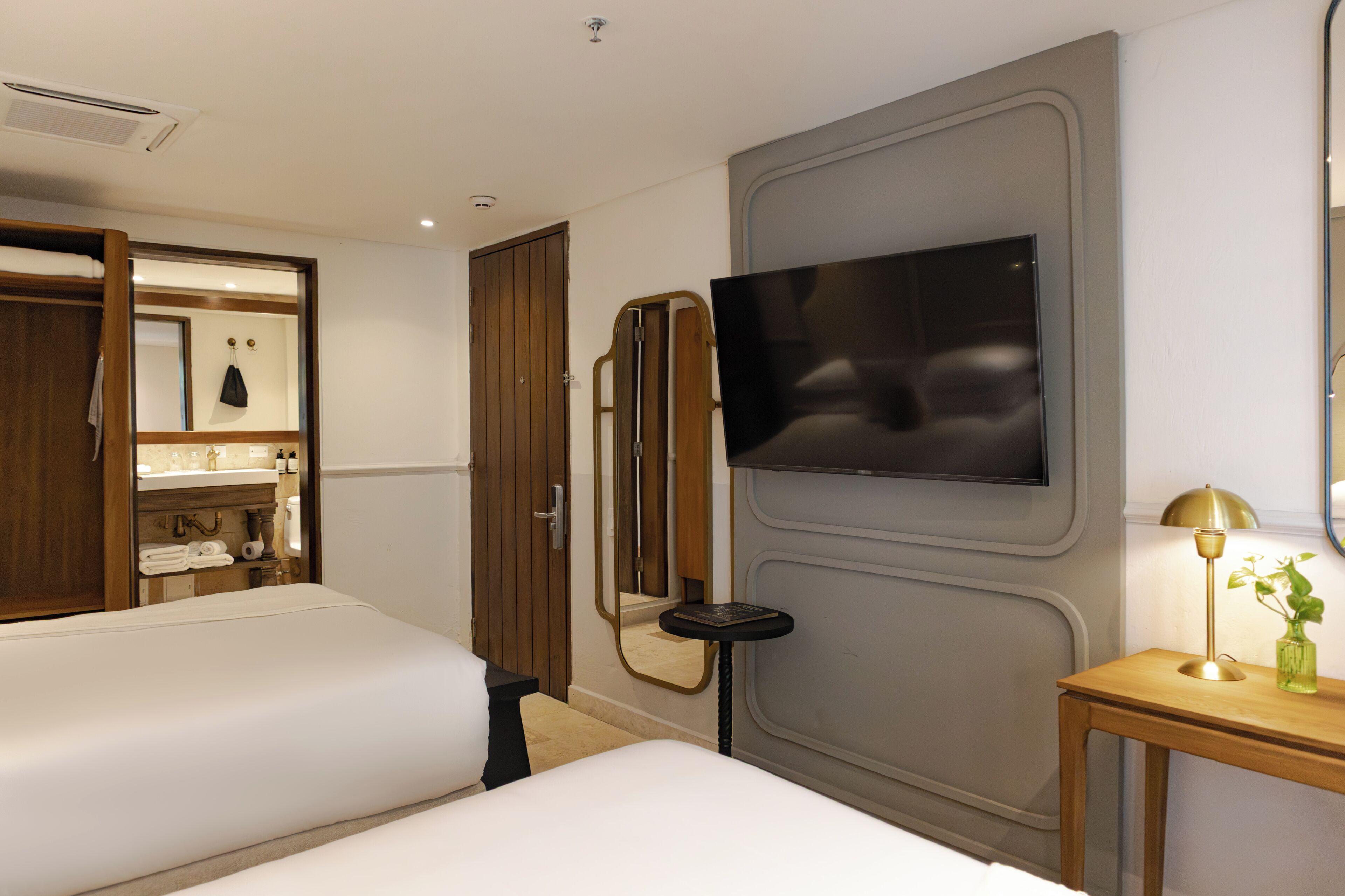 A hotel room with two beds, a TV, a mirror, a small table with a lamp, and a bathroom with a vanity and towel rack.