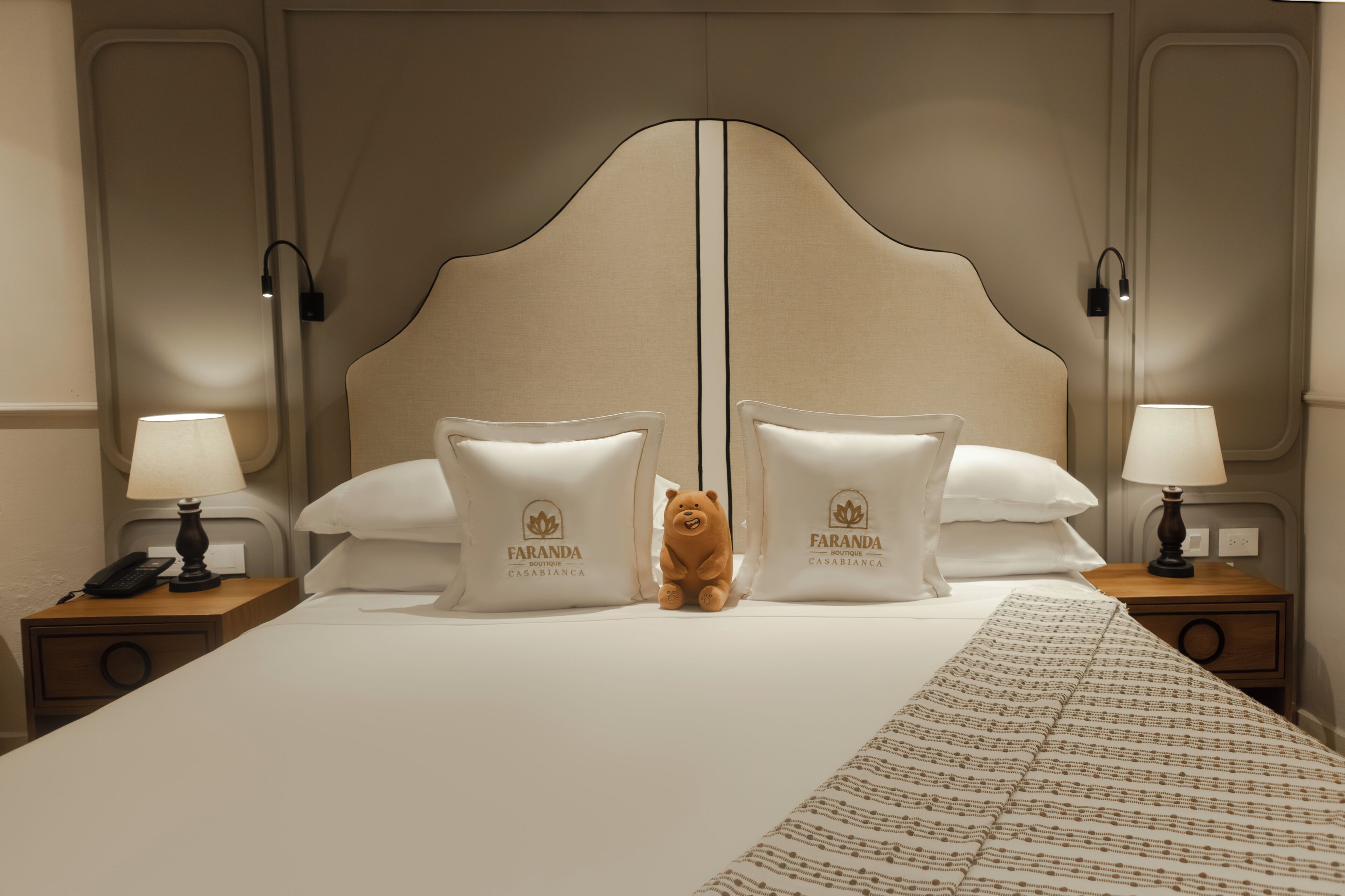 Habitación de hotel con una cama grande, dos mesitas de noche con lámparas, un oso de peluche y almohadas con el nombre del hotel.